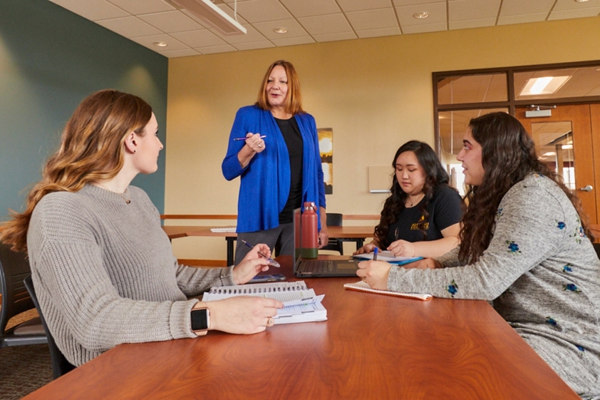 Group of three students sitting in a classroom at a table listening to professor teach
