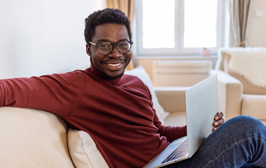 Black man in red shirt and glasses smiles while sitting on couch with a laptop