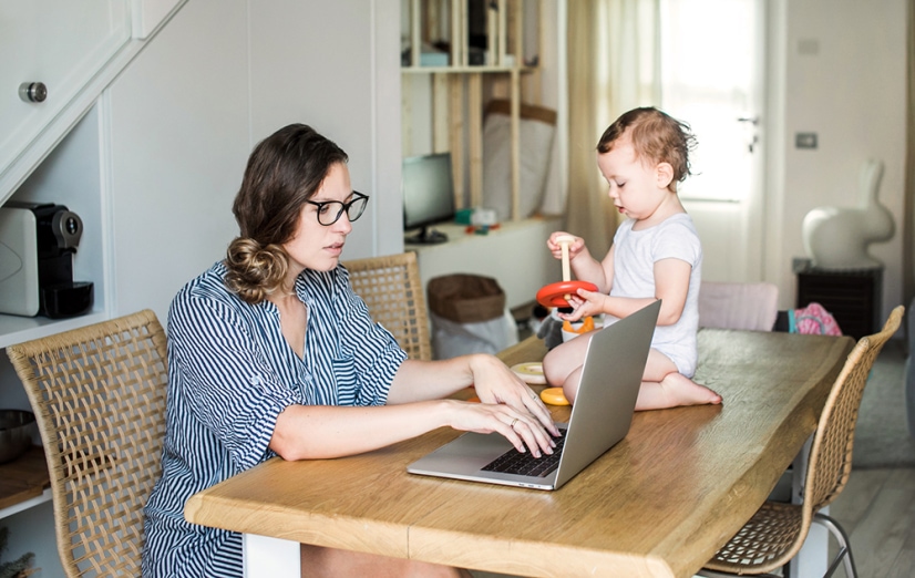 Young woman works on a laptop while her toddler sits next to her