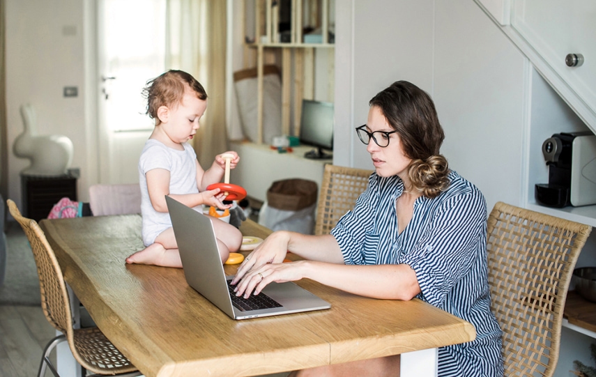Woman works on laptop on table with toddler next to her