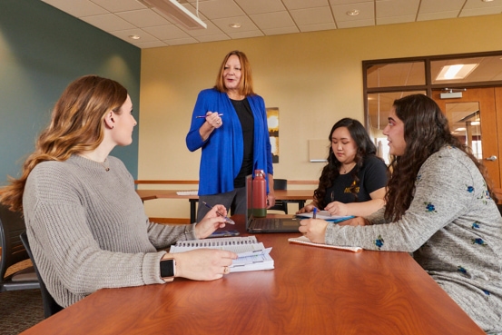 Group of three students sitting in a classroom at a table listening to professor teach