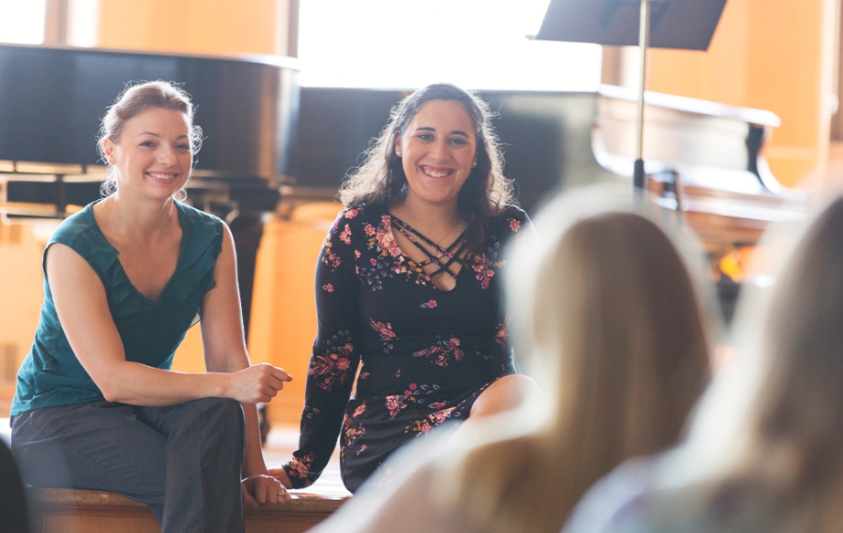 A music teacher and student sit on a stage in front of a grand piano while smiling to other students in the audience.