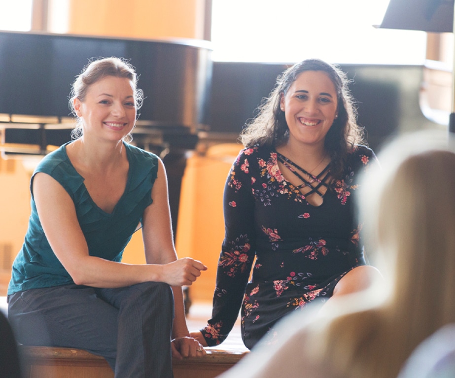 A music teacher and student sit on a stage in front of a grand piano while smiling to other students in the audience.